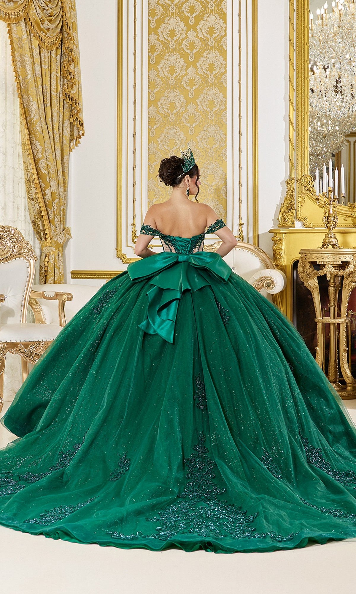 A woman wearing the Quinceanera Dress 5059 by Dancing Queen, featuring a large bow, stands with her back turned in an ornate room adorned with gold accents and patterned wallpaper.